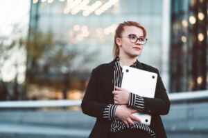 Junge Frau in Business-Kleidung mit Brille hält einen Laptop im Freien