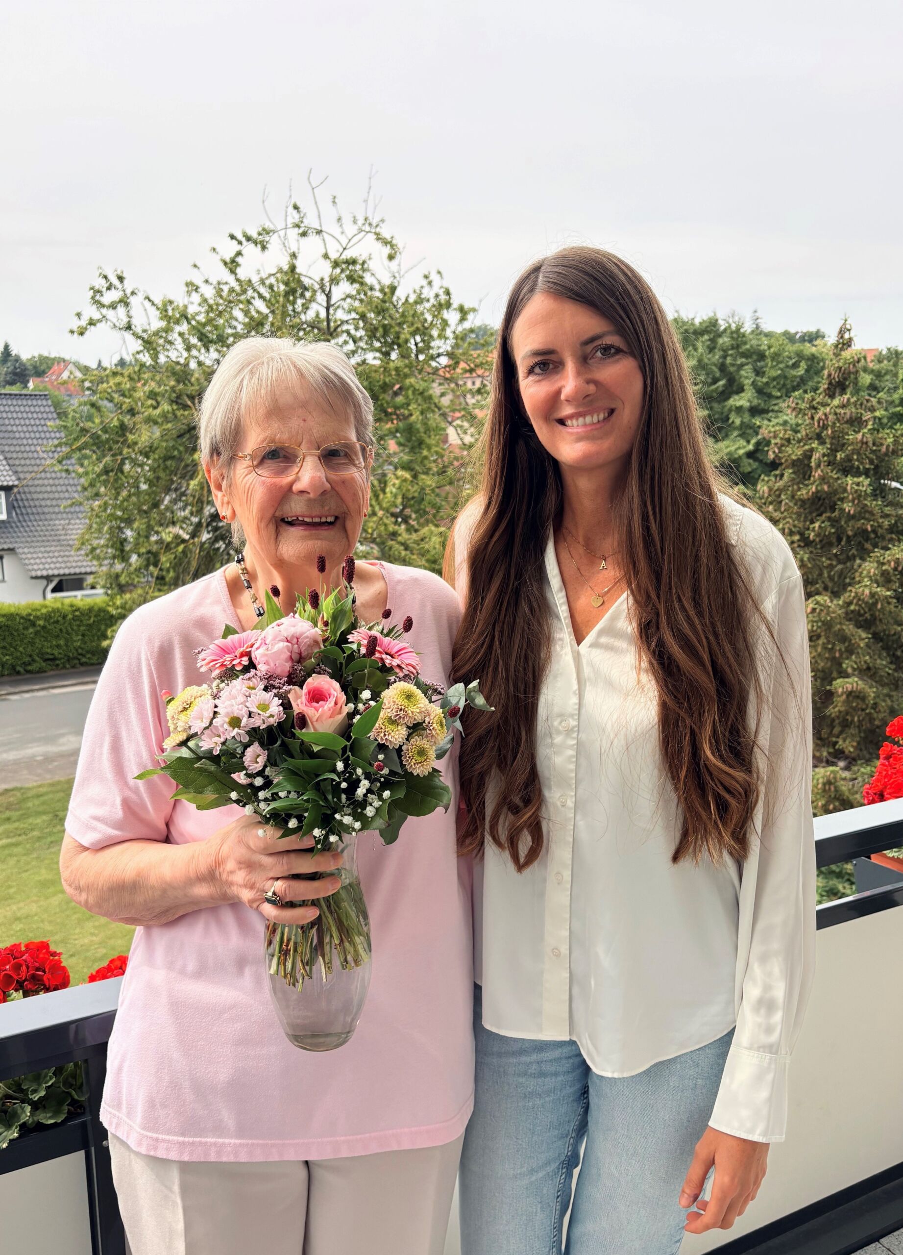 Zwei Frauen stehen auf einem Balkon, die linke Frau hält einen Blumenstrauß